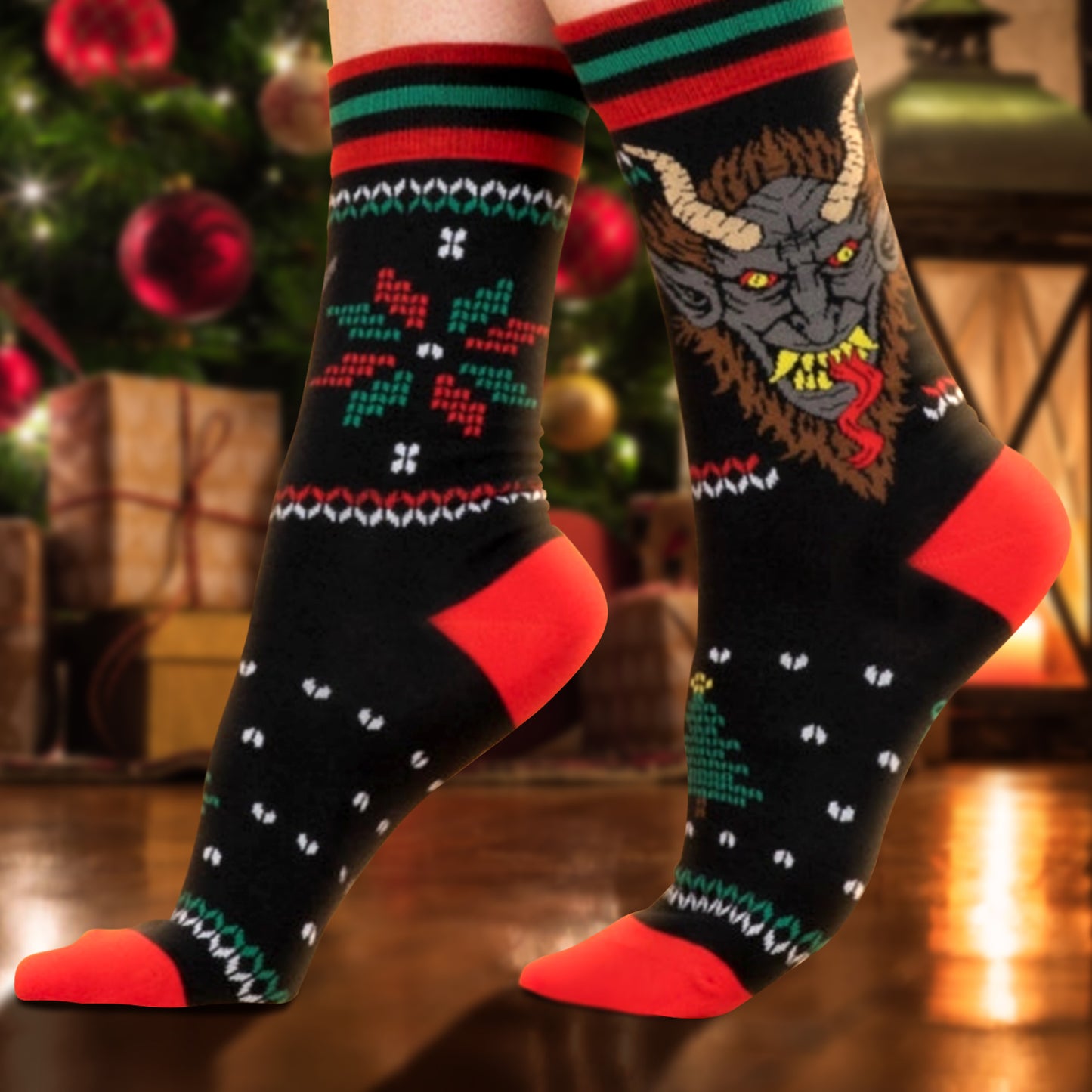 A model's feet wearing a pair of black socks with red and green stripes at the top. One side has a depiction of a red and green snowflake shape. The other side depicts the face of the mythological figure Krampus, with long horns on his head, sharp teeth, and a long red tongue. In the background is a Christmas tree next to an oil lamp.