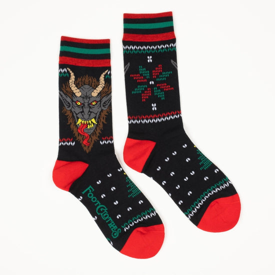 A pair of black socks with red and green stripes at the top. One side has a depiction of a red and green snowflake shape. The other side depicts the face of the mythological figure Krampus, with long horns on his head, sharp teeth, and a long red tongue.