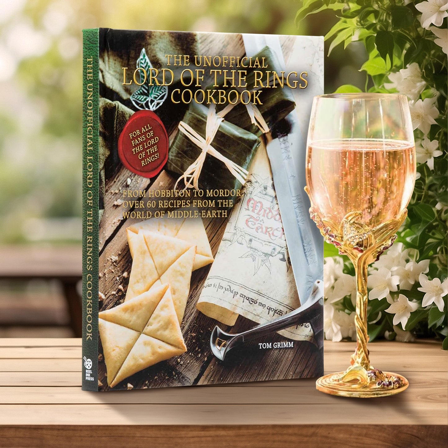 A copy of "The Unofficial Lord of the Rings Cookbook". The cover also reads "For all fans of Lord of the Rings" and "From Hobbiton to Mordor: Over 60 Recipes From the World of Middle-Earth - by Tom Grimm". The book is sitting beside a gold goblet/glass of a sparkling beverage.