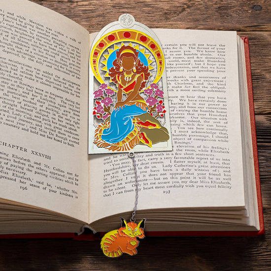 A metal bookmark set inside the pages of an open hardcover book. The bookmark depicts the character Leetah from the ElfQuest series. At the end of the bookmark is a chain with an orange and yellow charm, shaped like a cat