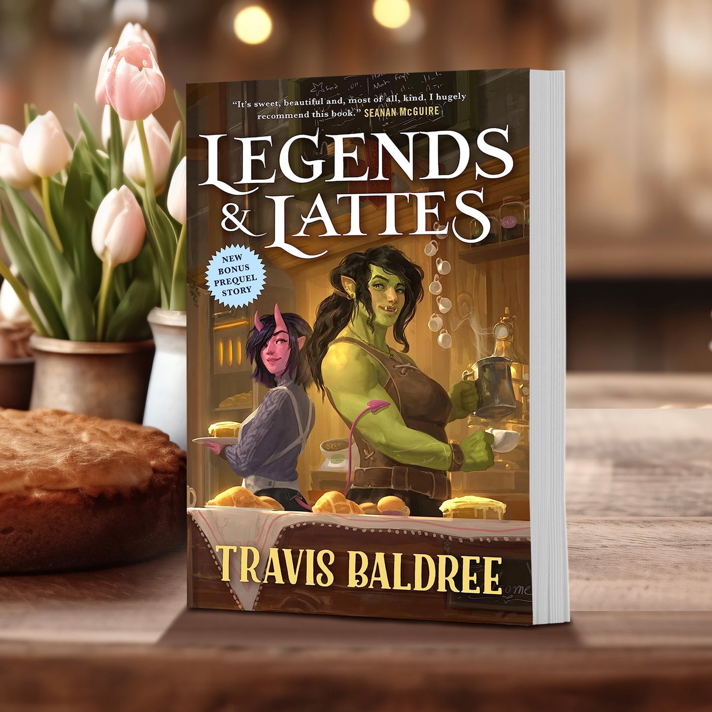 A book standing on a wooden kitchen counter. The cover depicts two female characters, one with green skin and one with pink skin, behind the counter of a coffee ship. At the top of the cover is white text that says "Legend and Lattes." At the bottom in yellow text is the author's name, Travel Baldree.