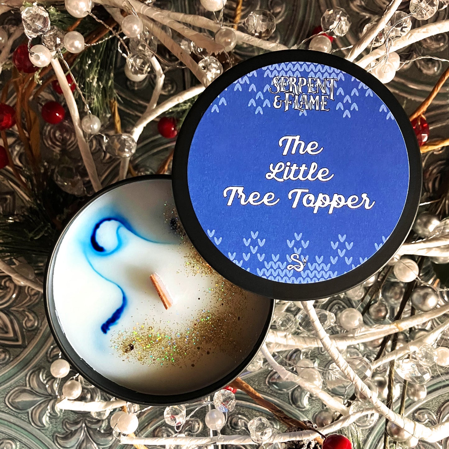 A black candle tin containing a white candle with a blue swirl and copper candle-safe glitter. The blue-and-white label reads "Serpent & Flame. The Little Tree Topper." The candle is sitting on a pile of white beads strung on white cords.