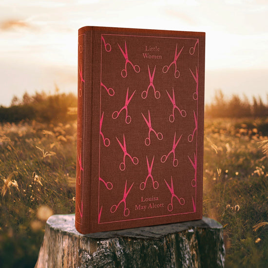 A clothbound hardcover copy of Louisa May Alcott's LITTLE WOMEN. It's a dark maroon cloth covered with a pattern of red scissors. The book is sitting on a tree stump in a field of flowers, with a bright sky in the background.