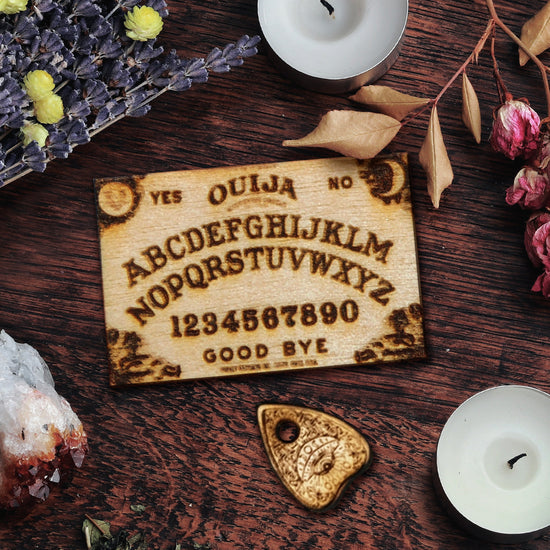 A small wooden wood-burned ouija board, with the traditional "yes/no", alphabet, numbers, and "good bye" in the Ouija layout. It's next to a tiny matching planchette and surrounded by witchy aesthetics.