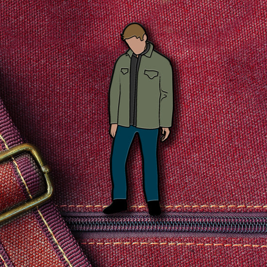 An enamel pin depicting Dean Winchester from the TV series "Supernatural." Dean is in a green jacket and blue pants and looking down at the ground. The pin is attached to a maroon canvas bag