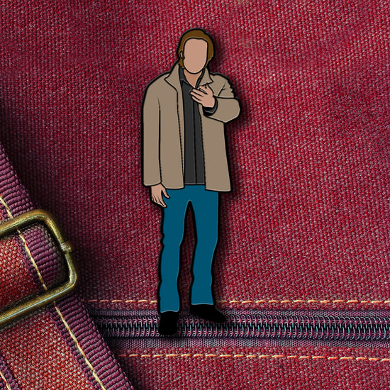 An enamel pin depicting Sam Winchester from the TV series "Supernatural." Sam is in a tan jacket and blue pants and holding his hand out. The pin is attached to a maroon canvas bag