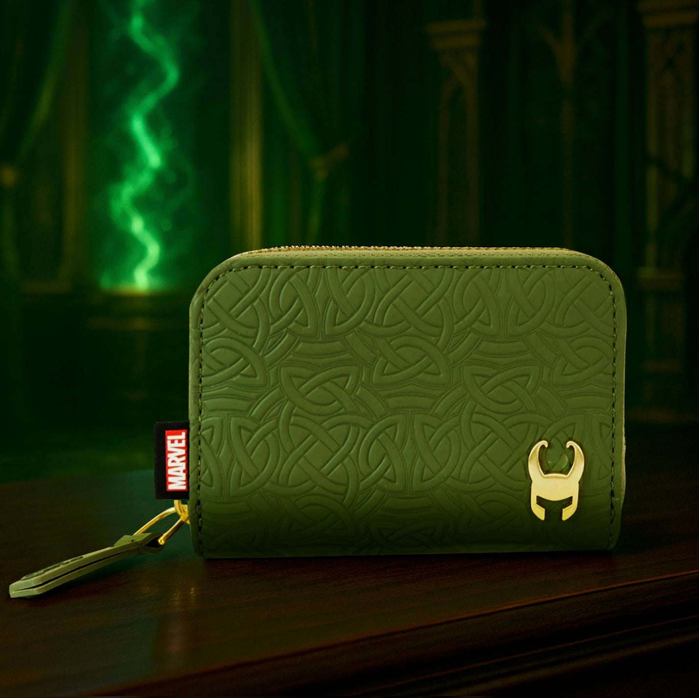 A green faux leather wallet with a zipper along the edge, on a dark wood table. On the front of the wallet is a repeating Norse knot pattern. On the bottom right of the wallet is a gold metal sticker representing the character Loki from the Marvel movie and TV series. In the background is a dark hall with a streak of green light in the corner.