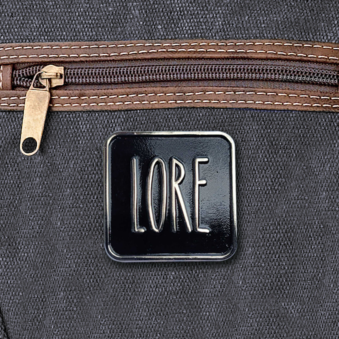 Load image into Gallery viewer, A silver enamel pin with a black rectangular front, which reads LORE in tall, skinny white font. The pin is affixed to a black canvas bag.
