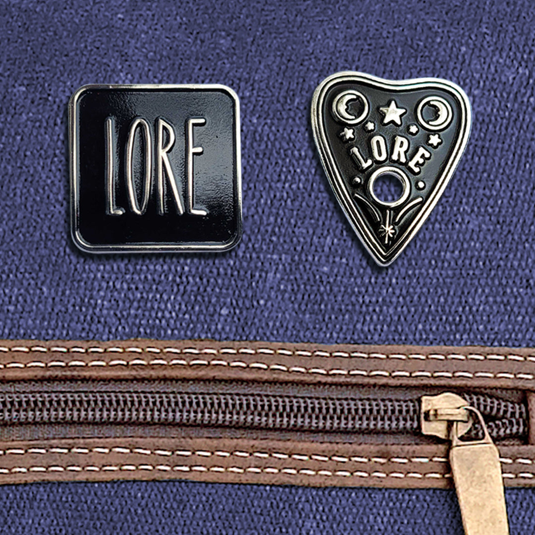 Two enamel pins side by side, attached to a blue canvas bag above a brown zipper. The left pin has a black rectangular front, which reads LORE in tall, skinny white font. The right pin has a triangular shape resembling a OUIJA planchette with stars and two crescent moons, which reads LORE in tall, skinny white font.