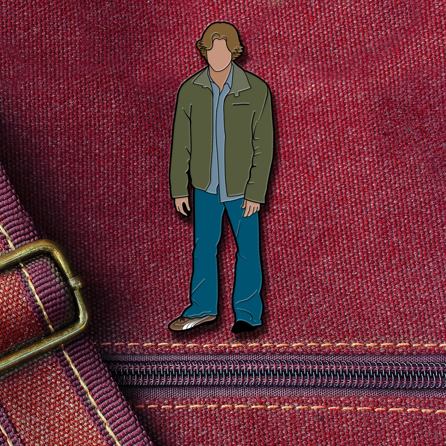 An enamel pin depicting Sam Winchester from the TV series "Supernatural." Sam is wearing jeans and a green jacket, and is missing his left shoe. The pin is attached to a maroon canvas bag.