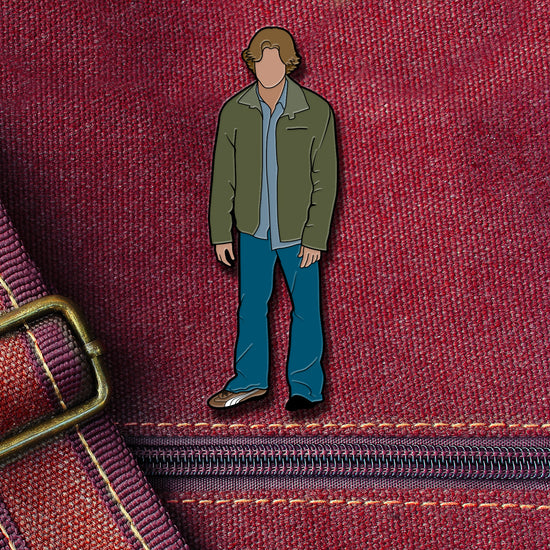 An enamel pin depicting Sam Winchester from the TV series "Supernatural." Sam is wearing jeans and a green jacket, and is missing his left shoe. The pin is attached to a maroon canvas bag.