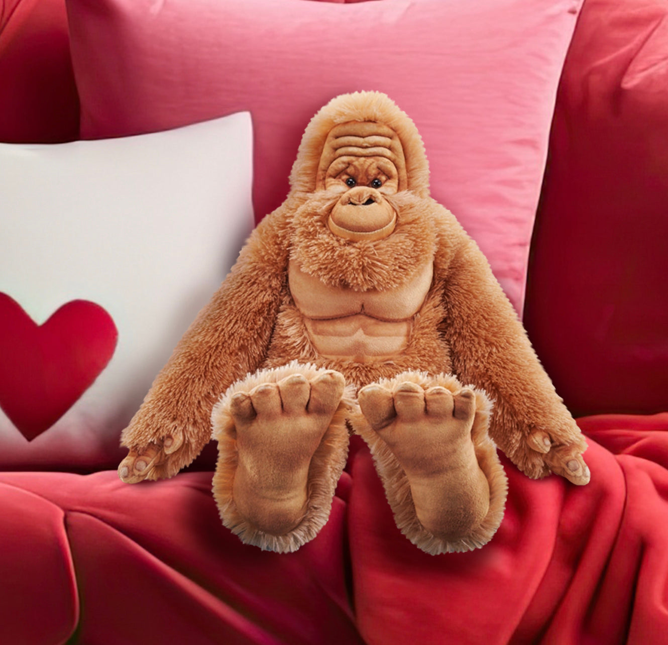 Close up view of a stuffed Bigfoot doll, "sitting" on a red-and-pink couch with throw pillows. The doll has reddish hair, with a light brown wrinkled face and very large brown feet.