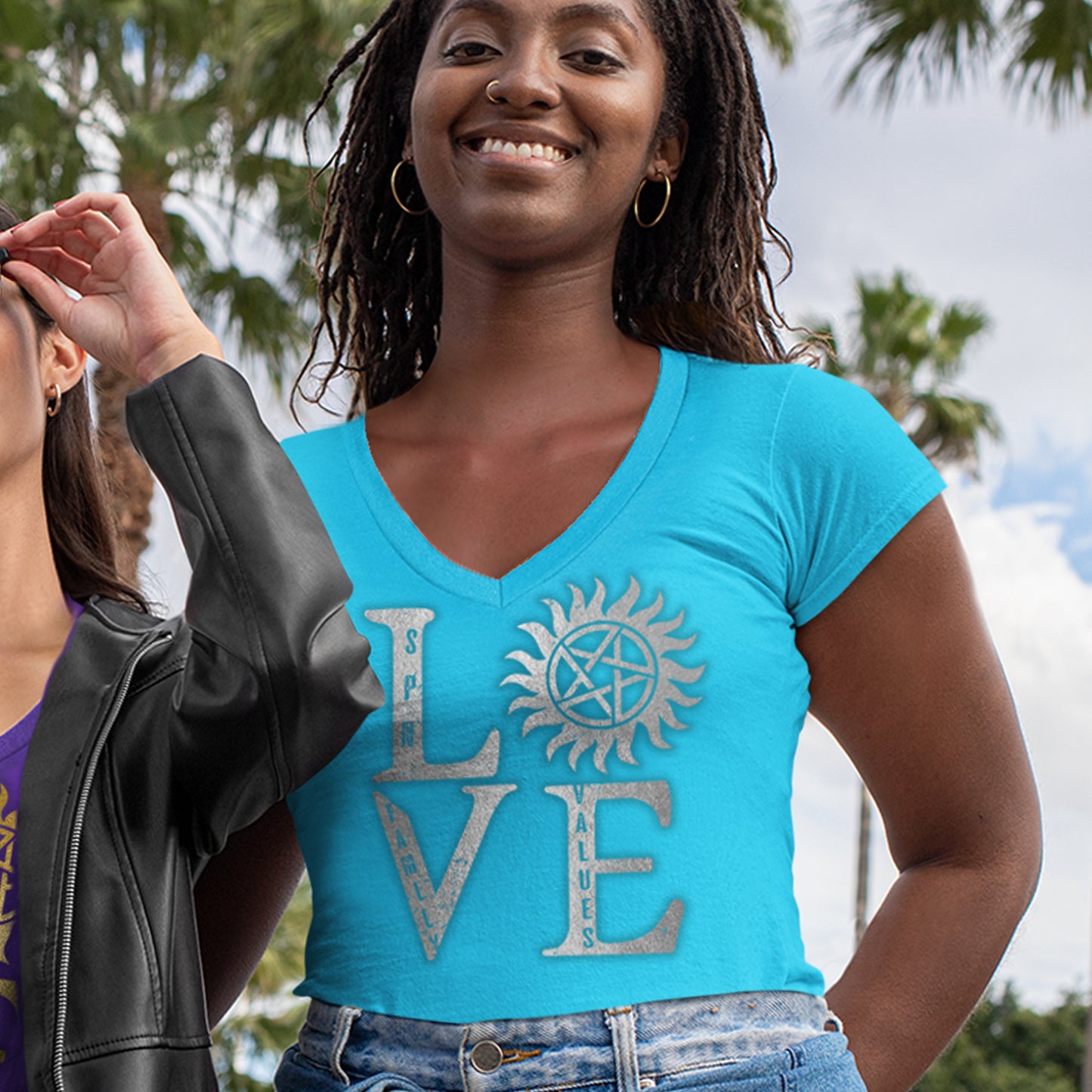 A female model wearing a turquoise v-neck shirt. The front of the shirt has silver text saying "love," with the "O" replaced by the anti-possession symbol. Next to the her is another model in a black leather jacket. In the background are palm trees and a cloudy sky.