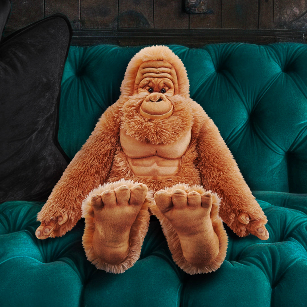 Close up view of a stuffed Bigfoot doll, on a teal green plush sofa. The doll has reddish hair, with a light brown wrinkled face and very large brown feet.