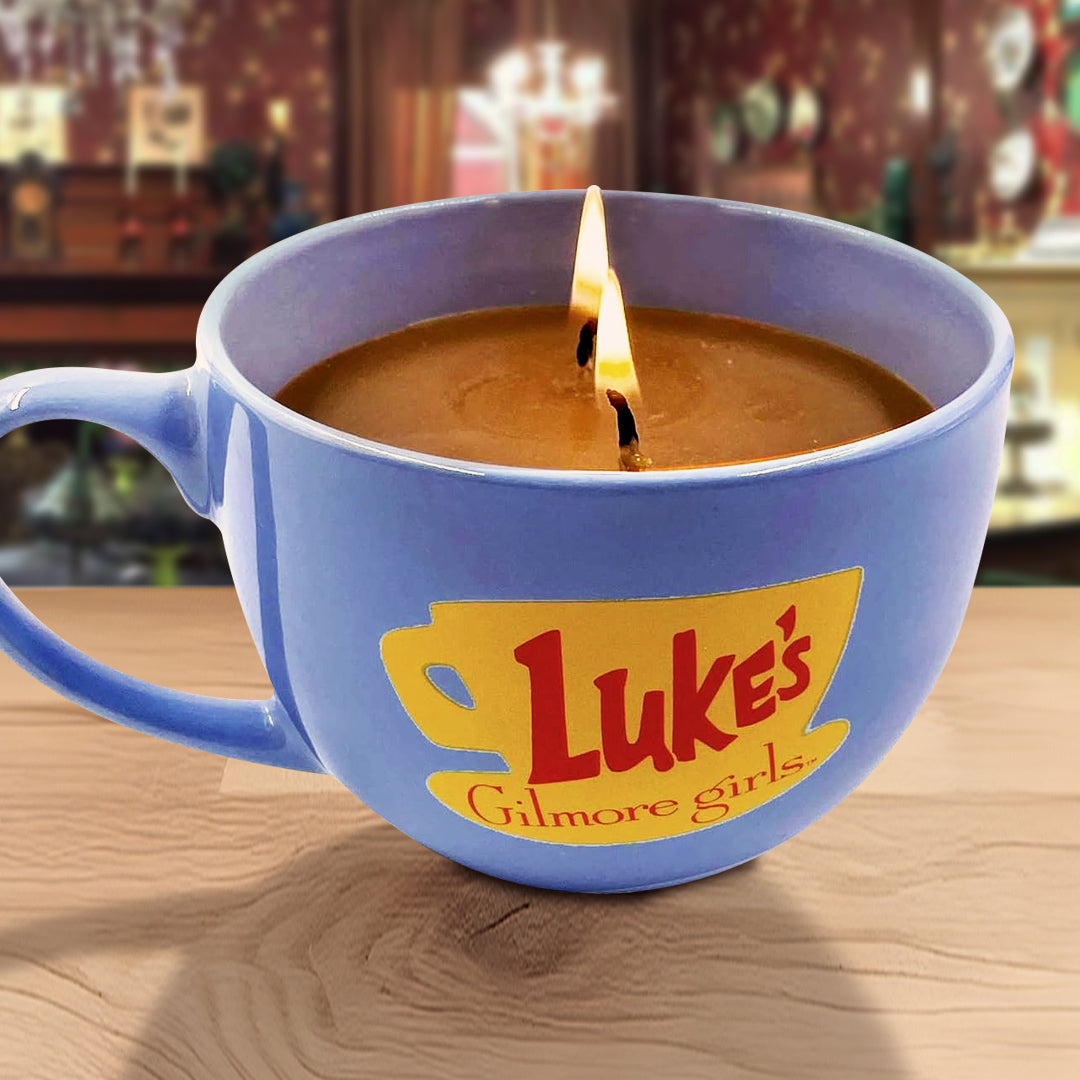 Blue mug with 'Luke's Diner' logo containing a candle on a wooden surface with the Dragonfly Inn in the background.