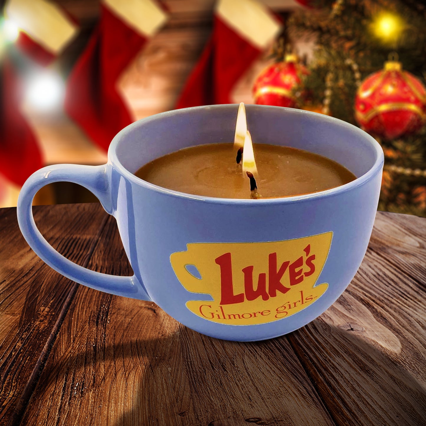Blue mug with 'Luke's Diner' logo containing a candle on a wooden surface with Christmas decorations in the background.