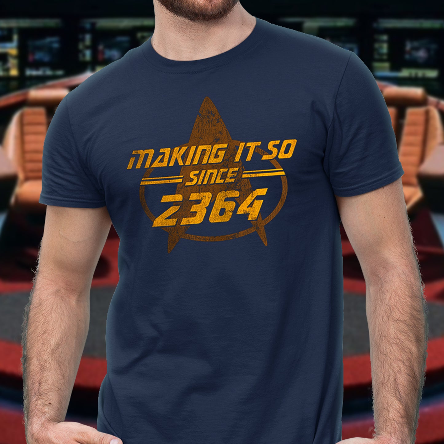 A man wearing a navy blue t-shirt with a distressed yellow graphic that reads 'making it so since 2364' inside a Star Trek-style delta emblem. The background is a blurred sci-fi corridor.