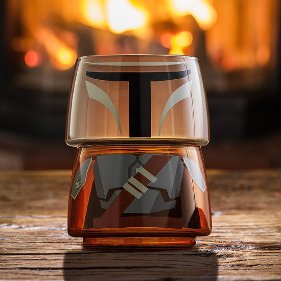 Two small drinking glasses stacked on each other on a wood table, with a fire in the background. The two glasses combined depict the titular character from the Star Wars series The Mandalorian.