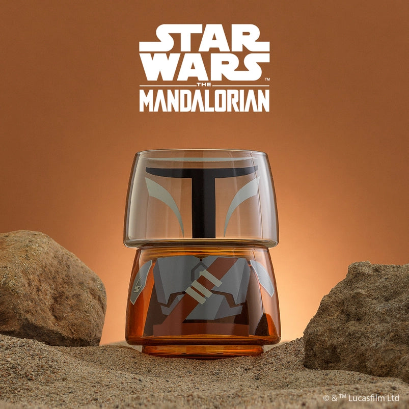 Two small drinking glasses stacked on each other on a stretch of sand, with a dark orange background behind them. The two glasses combined depict the titular character from the Star Wars series The Mandalorian. White text at the top says "Star Wars."