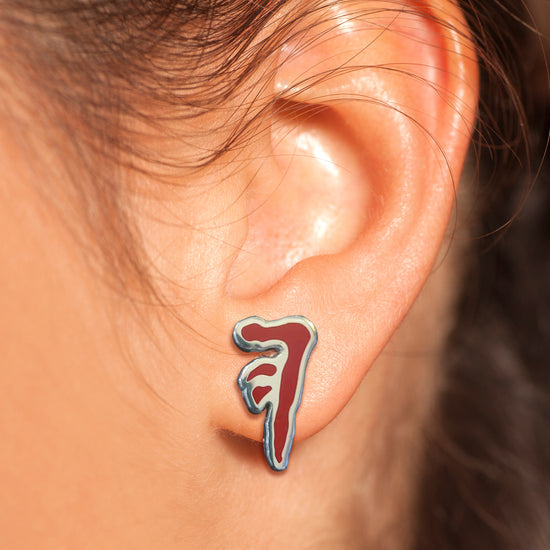 Close up of a model's ear, wearing an earring. The earring depicts the mark of cain in red and silver.