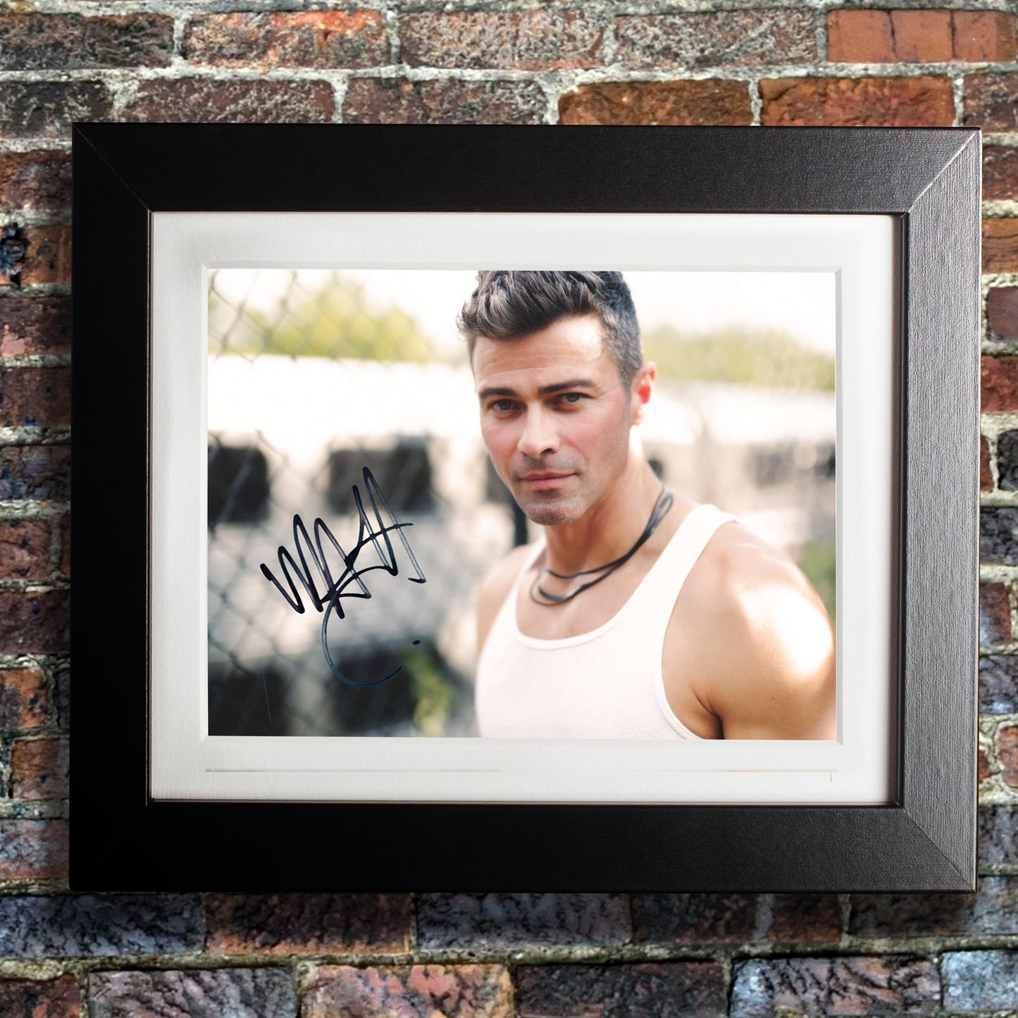 A black framed photograph against a brick wall. The photograph depicts actor Matt Cohen, dressed as a young John Winchester from the series "Supernatural." Matt is in a white tank top, standing in front of a chain link fence with a train in the background. On the left side is Matt's autograph in black ink.