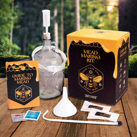 A black and yellow square box on a wood table, with a forest in the background. The top of the box depicts honey running down the sides. On the top of the box in yellow text is "mead making kit," with a drawing of a honeybee under it. Next to the box is glass jug. In front of the box mead-making tools that come with the kit.