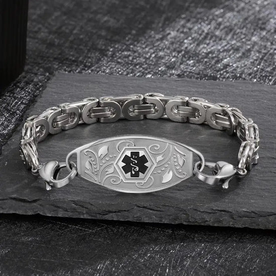 Close up view of a silver medical alert ID bracelet on a piece of black granite. The bracelet features the international medical symbol in the center, engraved in black. Around the symbol are raised depictions of leaves and flowers.