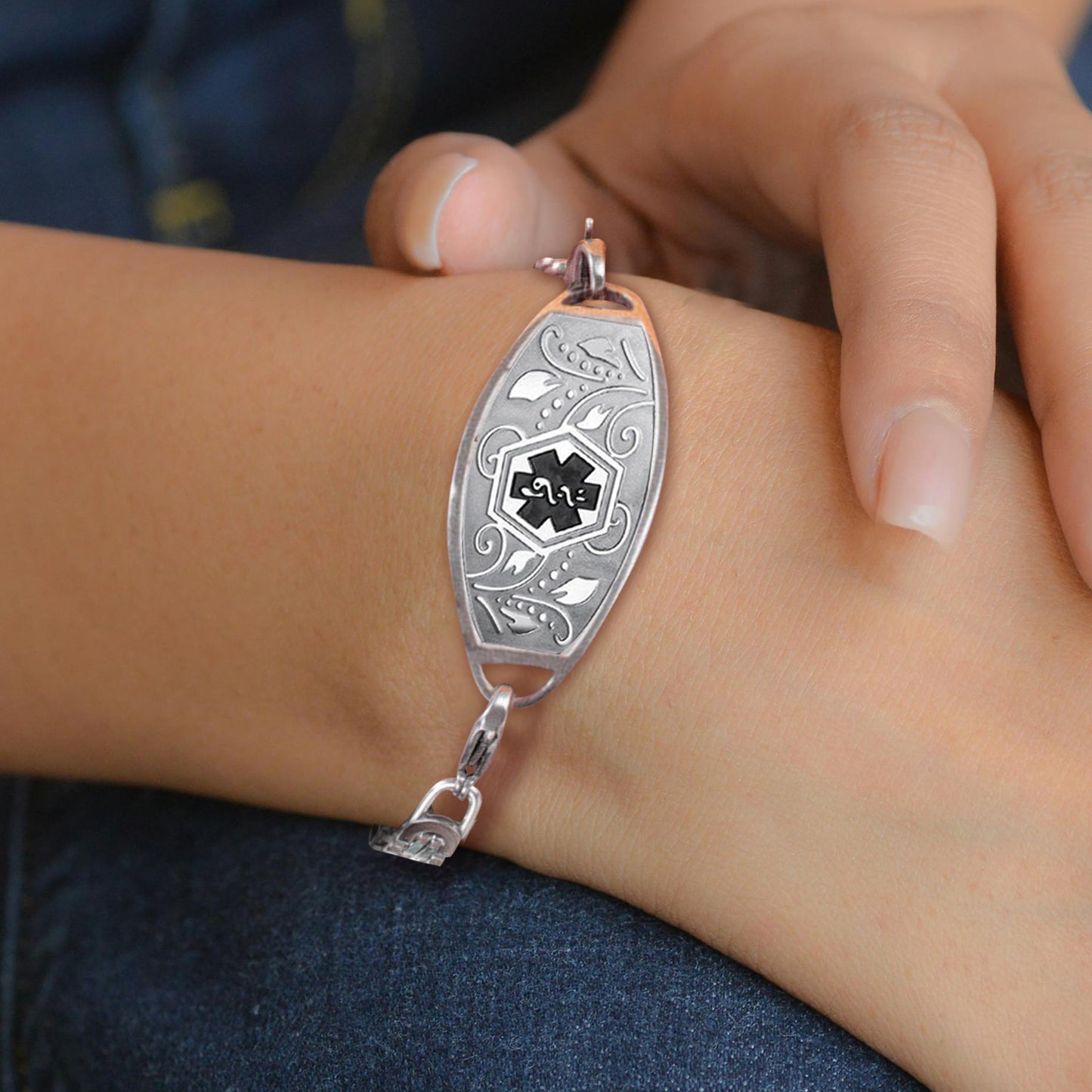 Close up of a model's hand with a silver Emergency Alert Bracelet around the wrist. The bracelet features the international medical symbol in the center, engraved in black. Around the symbol are raised depictions of leaves and flowers.