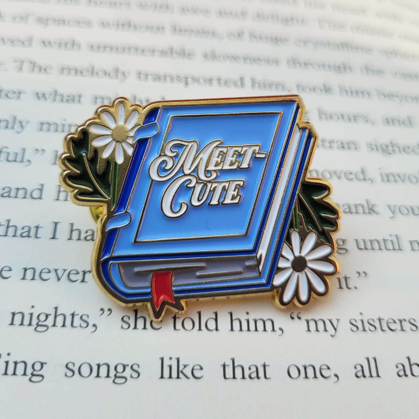 A blue enamel pin on a book page. The pin is shaped like a book, with white flowers on on each side, White text on the cover says "meet-cute."