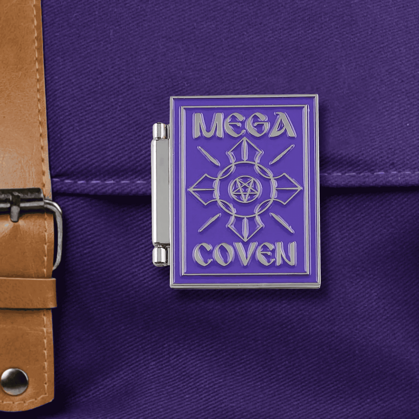 A silver enamel pin with a purple front in the shape of a book, with the words 'MEGA COVEN' in silver on the front. The hinge opens to reveal a spellbook page titled "World Domination Potion" on one side and a purple devil's trap on the other.