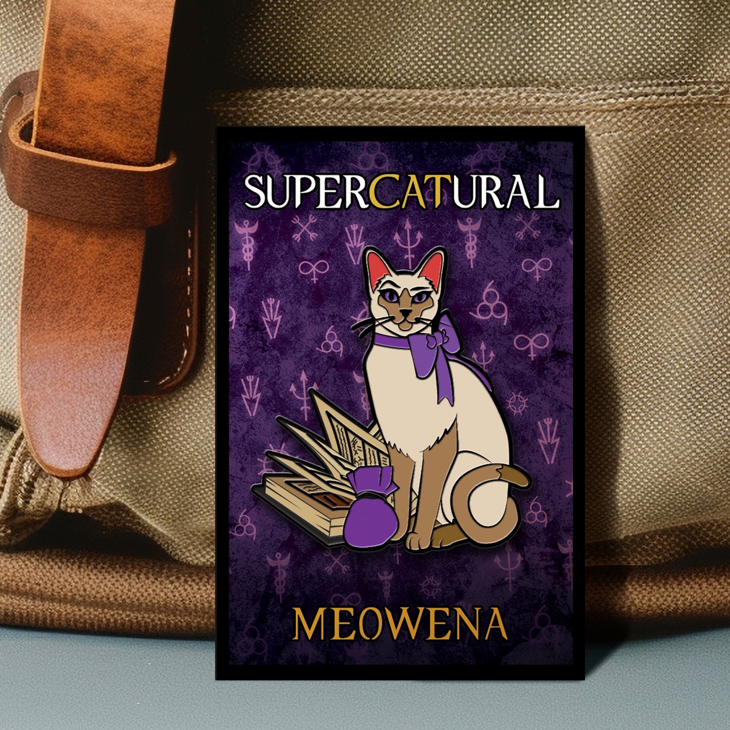 An enamel pin against a beige canvas bag. The pin depicts a Siamese cat with a purple bow around its neck. Next to the cat is a spell book, with a small purple bag in front of it. Behind the cat is a purple background with various magical symbols strewn across it. White and yellow text says "Supercatural: Meowena"