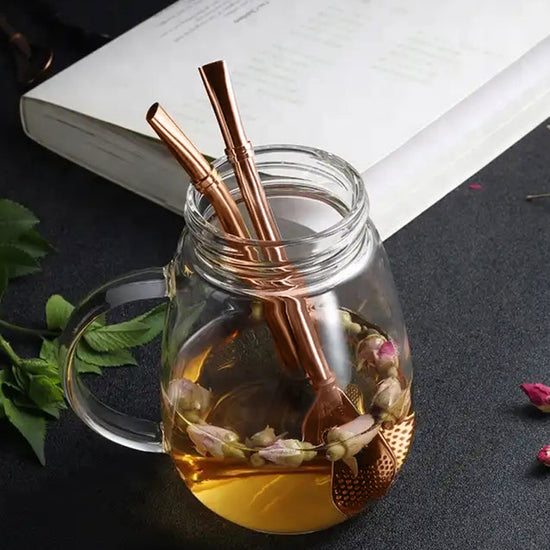 Close up view of a glass mug, partly filled with tea. Two metal drinking straws are inserted into the mug. Behind the mug is a white notebook on a black table top.