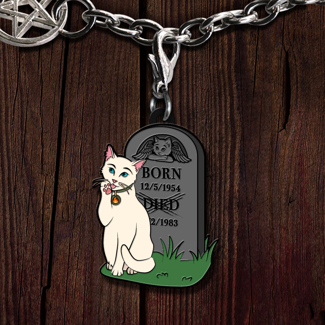 A close-up of a silver chain necklace with a custom engraved tombstone pendant featuring a white cat illustration and memorial text, set against a dark wood background.