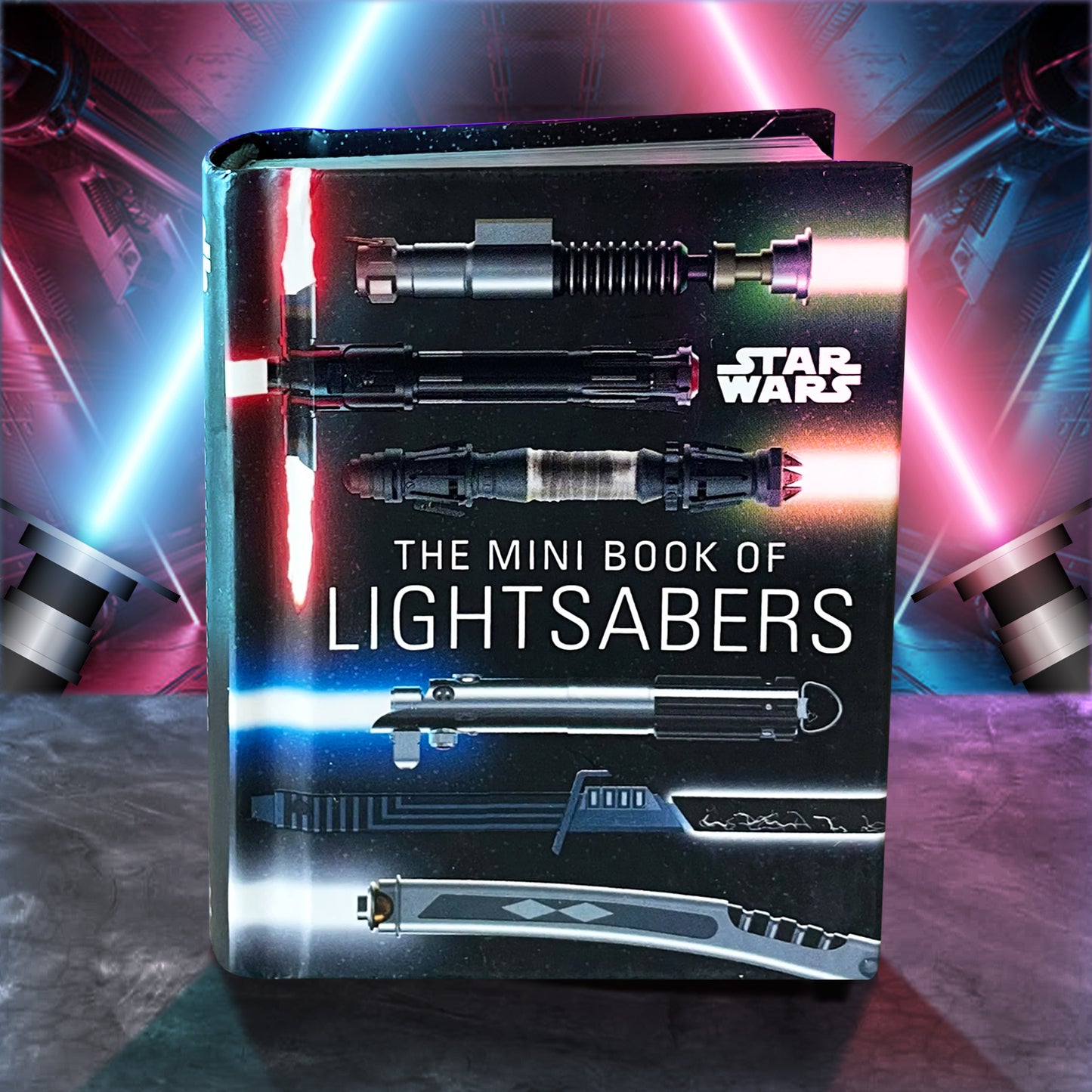 A small hardcover book titled "STAR WARS: The Mini Book of Lightsabers", with several lightstabers laying horizontally across the starry landscape. The background is a blue and red lightsaber in a dark ship. 