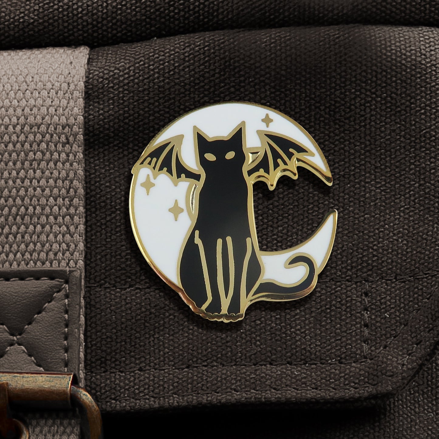 A black and white enamel pin with gold edges, attached to a black canvas bag. The pin depicts a black cat with bat wings, sitting upright. Behind the cat is a white crescent moon, with tiny gold stars scattered around it.