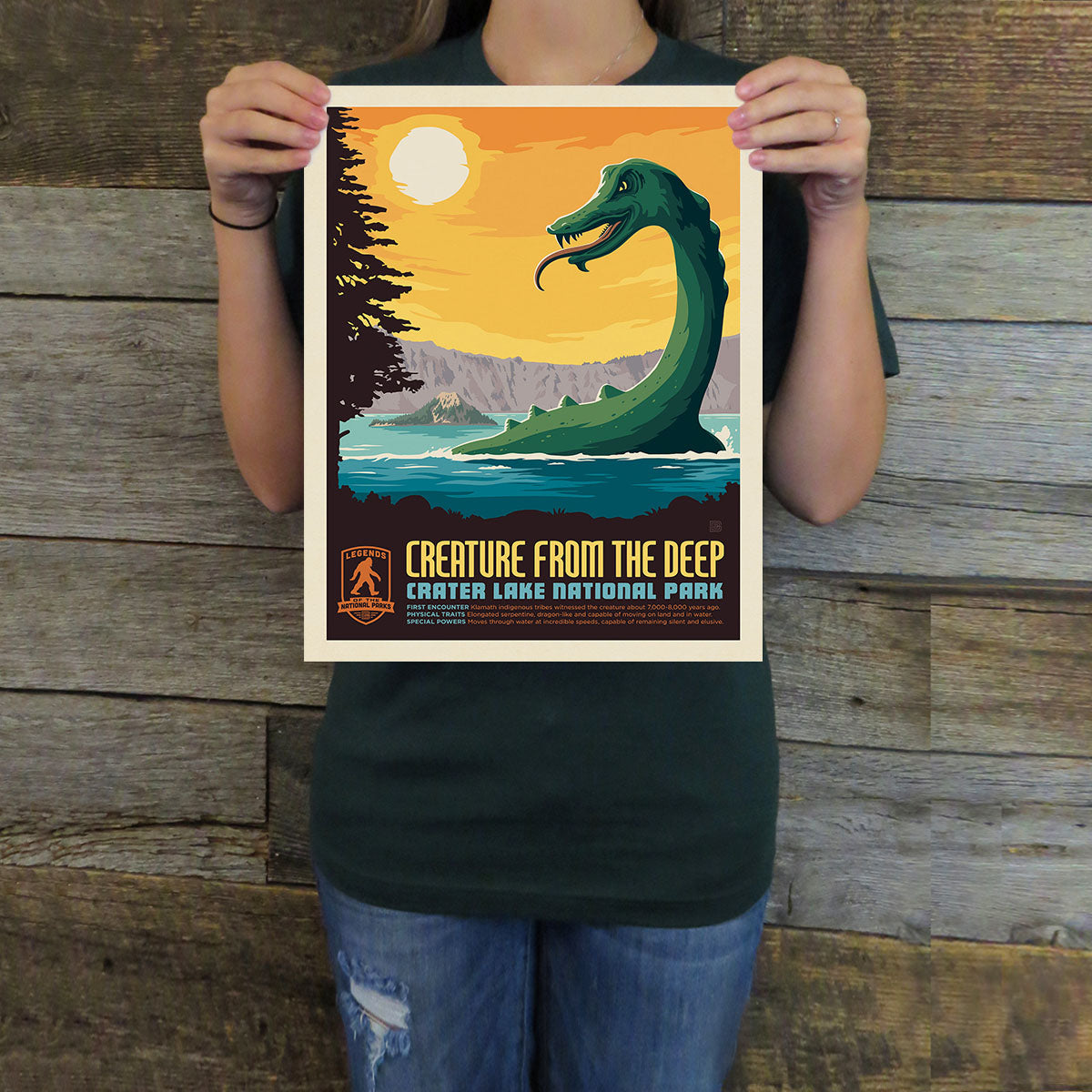 Person holding a poster of 'Creature from the Deep' at Crater Lake National Park against a wooden background