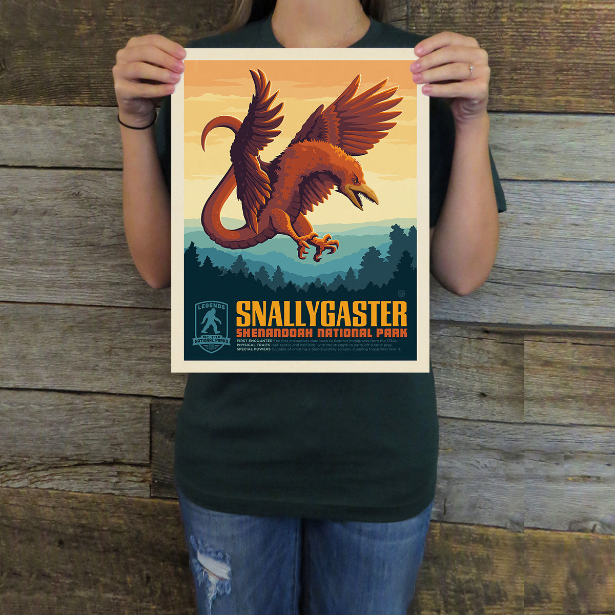 Person holding a poster of a dragon with 'Snallygaster' text against a wooden background