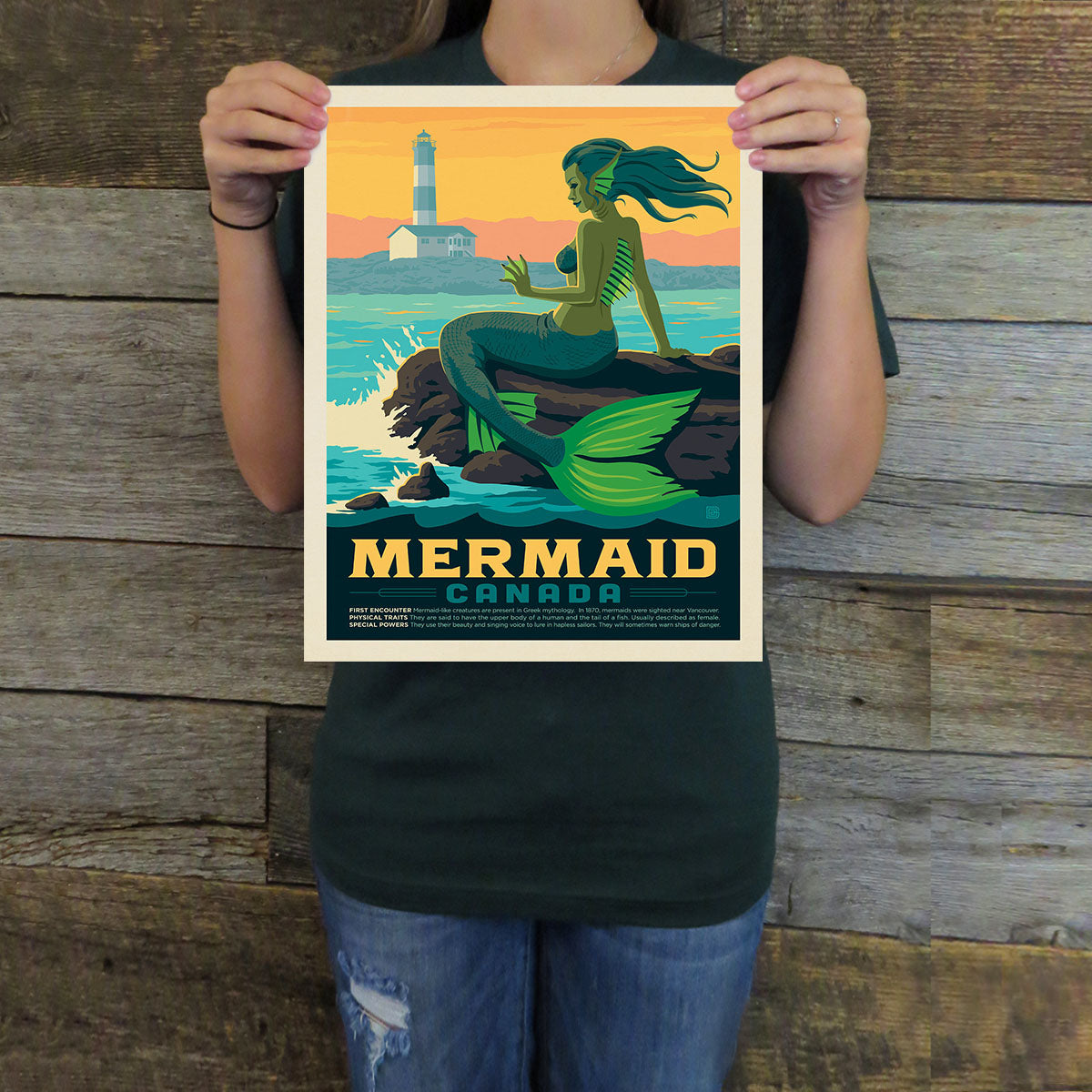 Person holding a poster of a mermaid with a lighthouse in the background against a wooden wall.