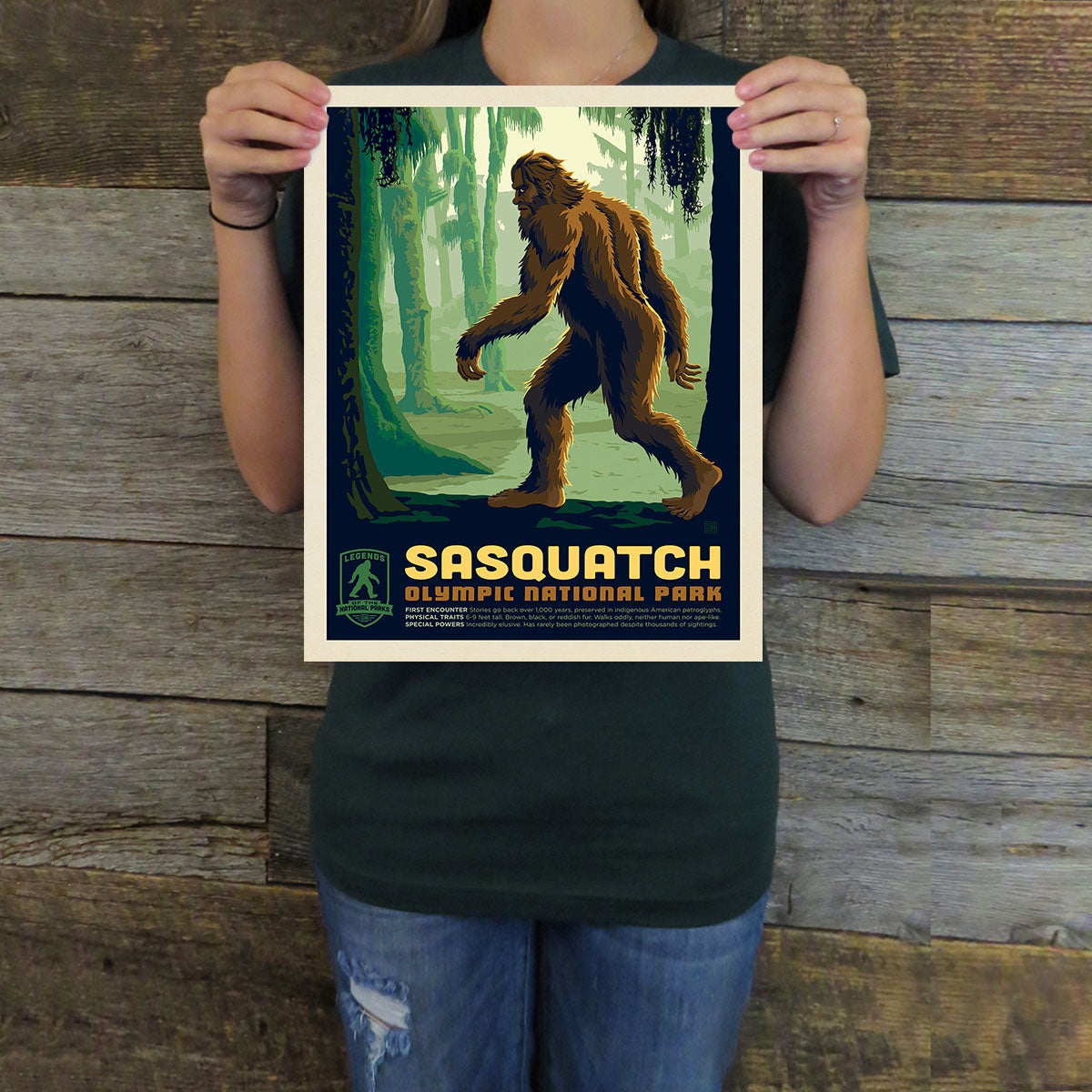 Person holding a Sasquatch Olympic National Park poster against a wooden background