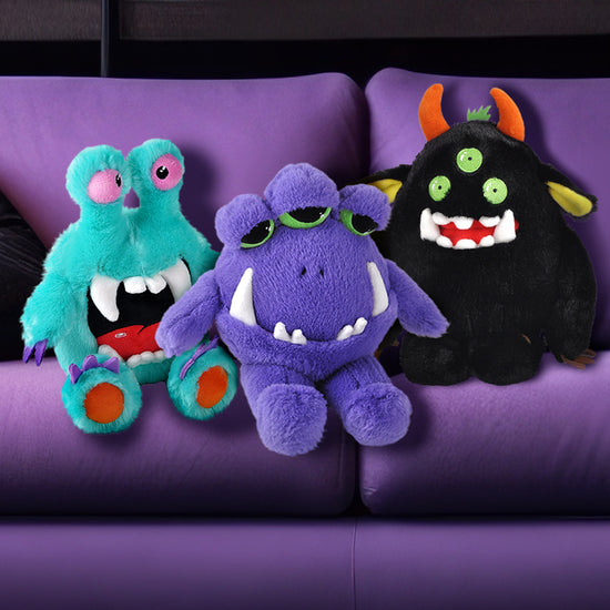 Three plushies sitting on a purple couch. One is blue with eye stalks on the top of his head. The second is purple with three half-opened green eyes. The third is black with three green eyes and orang horns. Each plushie has large white fangs sticking out of its mouth.