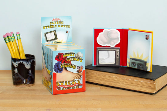 A light blue book of sticky notes on a wooden desk. On the cover is a depiction of the Monty Python's Flying Circus logo.Next to the book are sticky notes from the collection, deaturing images from the iconic TV series.  A black jar filled with pencils is next to the notes.