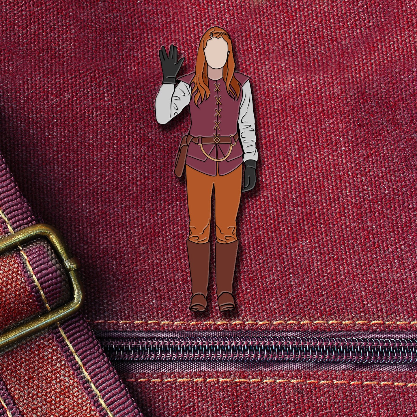 An enamel pin depicting Charlie Bradbury from the TV series "Supernatural." Charlie is wearing a medieval outfit, and holding one hand up in the Vulcan salute from Star Trek. The pin is attached to a maroon canvas bag
