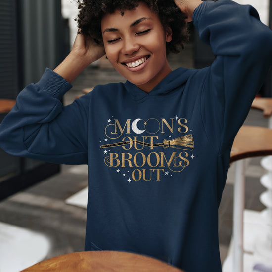 A female model wearing a navy blue hoodie with matching drawstrings, a kangaroo pocket, and a gold print reading "Moon's Out Brooms Out". There are a gold broom going through the first "Out" and small white stars dotting the print space. The first "O" in "Moon" contains a white crescent moon.  Behind the model is a cafe interior.