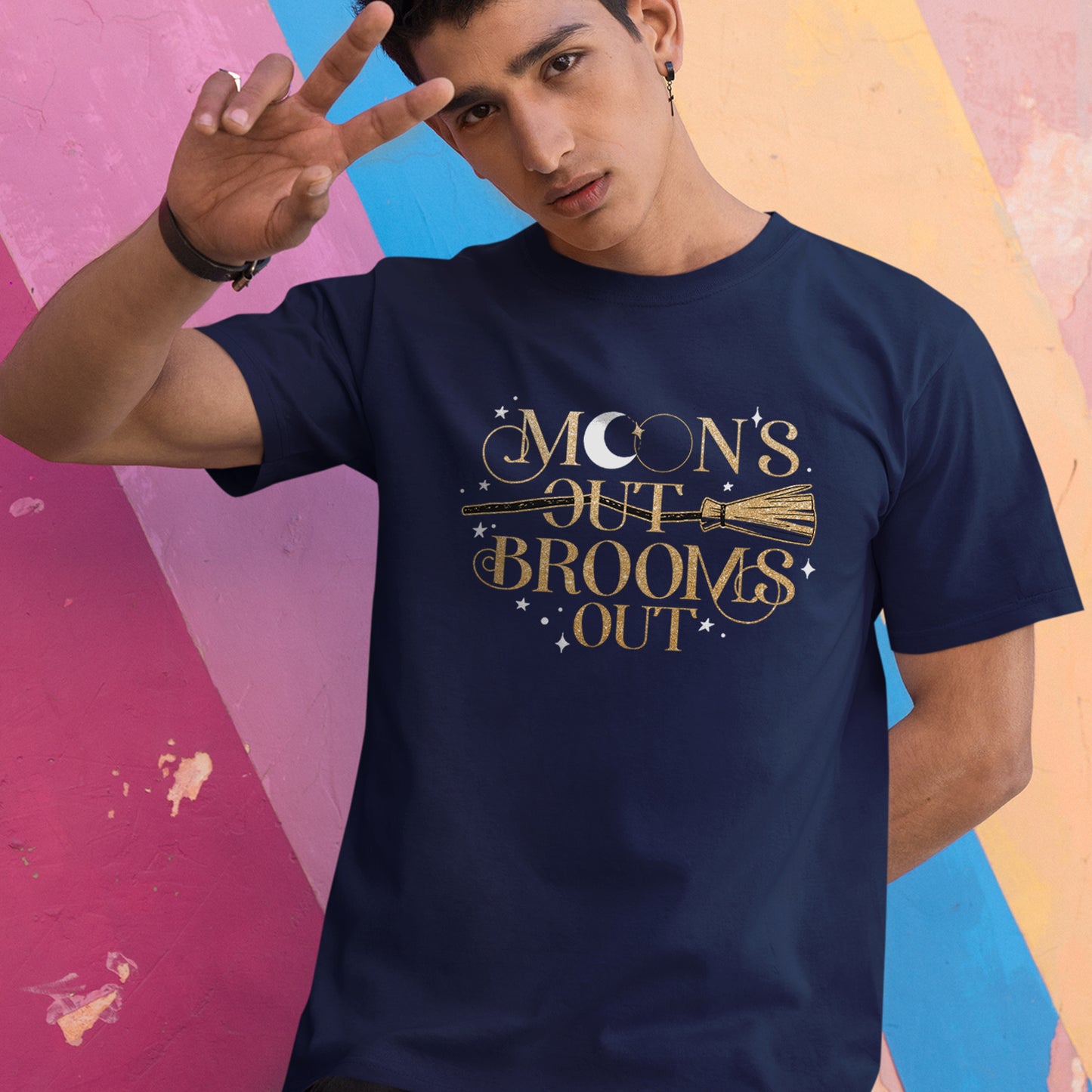 A male model wearing a dark teal, short-sleeved, crewneck tee with a gold print reading "Moon's Out Brooms Out". There are a gold broom going through the first "Out" and small white stars dotting the print space. The first "O" in "Moon" contains a white crescent moon.  Behind the model is a striped wall in rainbow colors.