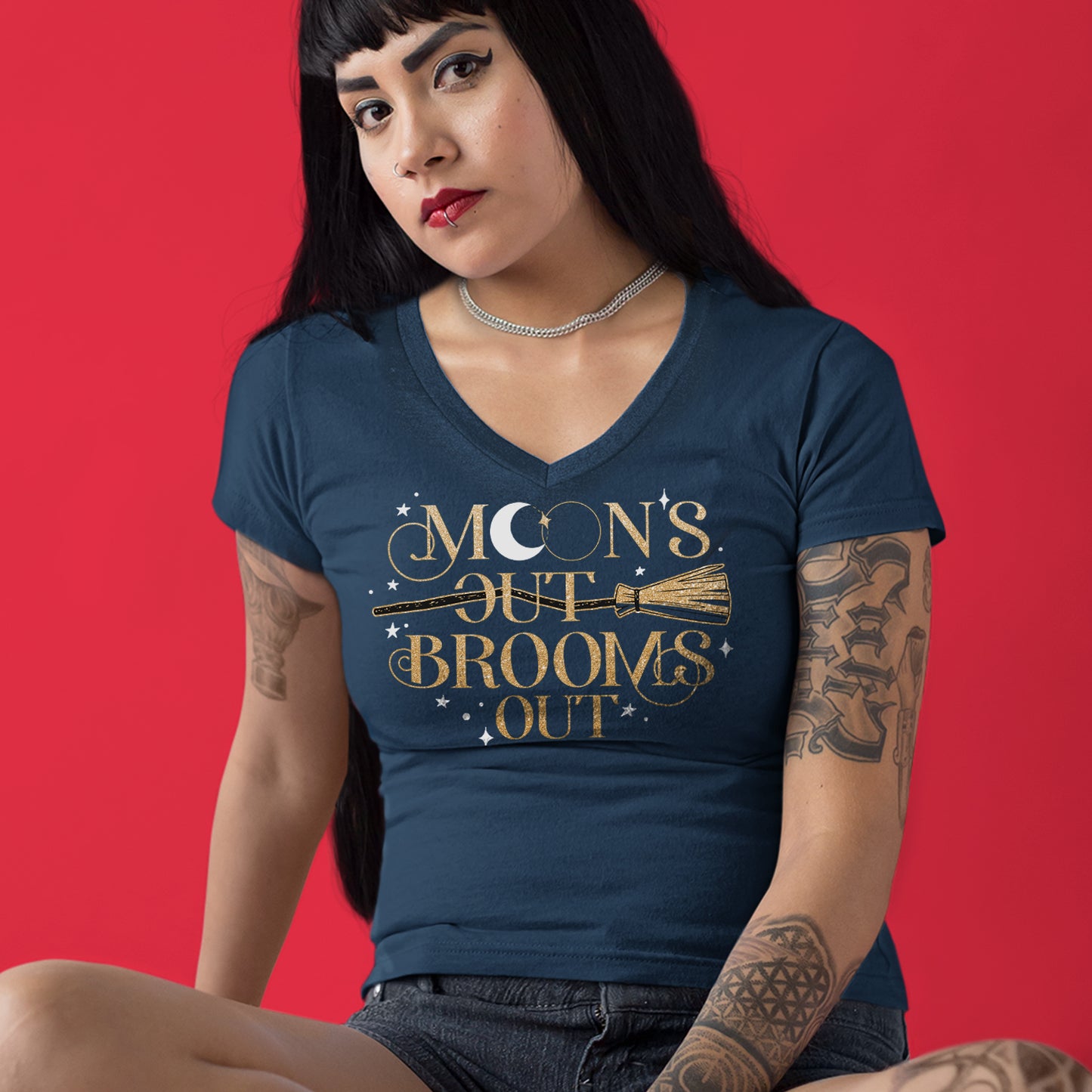 A female model wearing a dark navy, short-sleeved, V-neck tee with a gold print reading "Moon's Out Brooms Out". There is a gold broom going through the first "Out" and small white stars dotting the print space. The first "O" in "Moon" contains a white crescent moon. Behind the model is a red background.