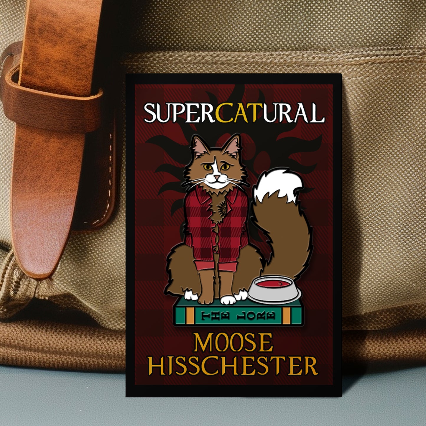 A rectangular enamel pin against a tan canvas bag. The pin depicts a Maine Coon cat wearing a red and black plaid shirt, sitting on a green boo entitled "The Lore." Next to the cat is a food bowl. Behind the cat is a red plaid background with a black paw print. White and yellow text says "Supercatural. Moose Hisschester"