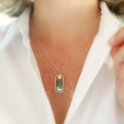 A rectangular pendant with rounded edges, against a model's neck. Inside the pendant is a depiction of a moose walking on front of pine trees, with mountains in the back, and a round golden sun at the top. A stainless steel necklace chain is attached to the pendant.