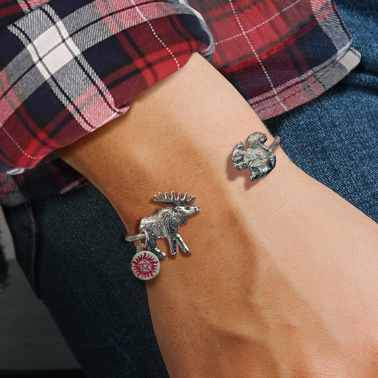 Close up view of a model's arm, with a bracelet  around the wrist. A moose bangle is attached to one end of the bracelet with an anti-possession charm under it. A squirrel bangle is at the other. The model is wearing a red, black, and white plaid shirt.