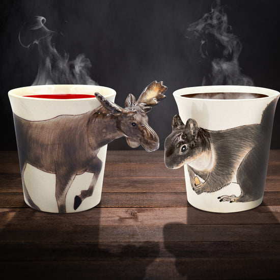 A pair of ceramic mugs on a wooden table. The left mug depicts a moose drawn on the front, with the moose's head sticking out as the mug handle. The right mug depicts a squirrel drawn on the front, with the squirrel's head sticking out as the mug handle. The Moose mug is filled with a red liquid, the squirrel mug is filled with coffee. Steam is rising from both mugs.