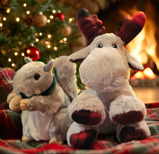 Two plushies on a red and green plaid blanket, in front of a Christmas tree next to a fireplace. The left plushie is a grey squirrel with a green plaid collar around his neck, holding an acorn between his paws. The right plushie is a grey moose with red and black plaid antlers and feet.